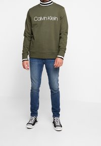 Felpa verde oliva con logo bianco "Calvin Klein", abbinata a jeans blu e sneakers nere. Presenta colletto e polsini a righe.