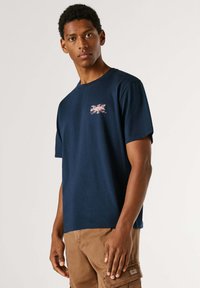 Young man in navy blue t-shirt with a small Union Jack design, wearing light brown cargo pants, standing against a plain background.