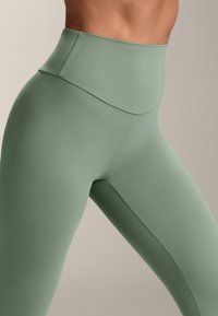 Visoke tajice u umirujućoj zelenoj boji, izrađene od rastezljivog materijala. Imaju glatku teksturu s čistim, minimalističkim dizajnom.