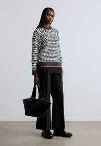 Svartvit mönstrad tröja med rosa kant, kombinerad med svarta byxor med vida ben och svarta läderskor. Modellen håller en svart handväska.