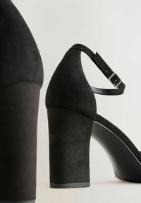 Sandale cu toc din suede negru, cu un toc chunky. Dispune de o curea la gleznă cu o cataramă argintie și o placă de bază netedă neagră. Suprafață superioară texturată.