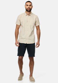 Beige kortærmet skjorte med knaplukning og brystlomme, parret med mørke rullede denimshorts og beige slip-on sko.