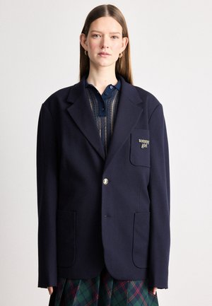 Blazer bleu marine surdimensionné avec deux poches avant, une fermeture à un bouton et des accessoires en or ; présente un patch logo sur le côté gauche.