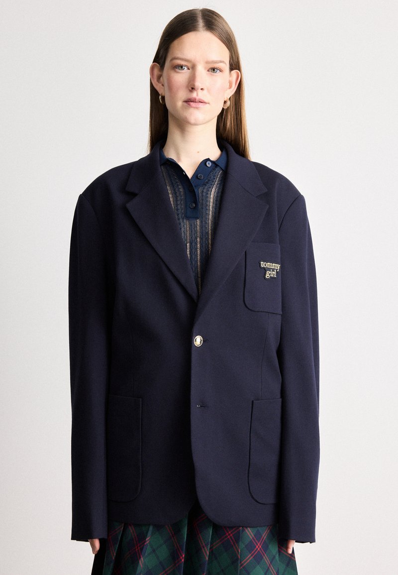 Blazer overtagliato navy con due tasche frontali, chiusura a un bottone e dettagli in oro; presenta una toppa con logo sul lato sinistro.