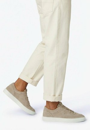 Persona che indossa sneaker in camoscio beige e pantaloni crema risvoltati, in piedi con un piede leggermente sollevato.