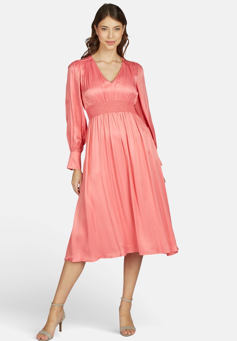 Vrouw in een koraalroze satijnen midi-jurk met lange mouwen en een smocktail, met beige hoge hakken aan, staand met één been naar voren.