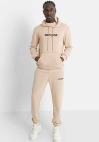 Conjunto de sudadera con capucha y pantalones deportivos beige con el logo "SIXTH JUNE" en negro, que incluye un bolsillo tipo canguro, cordones y puños elásticos. Zapatillas blancas.