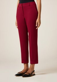 Kvinde iført bordeauxfarvede skræddersyede cropped bukser med ophøjede kanter og sorte spidse slingback flade sko med brune stribede stropper.