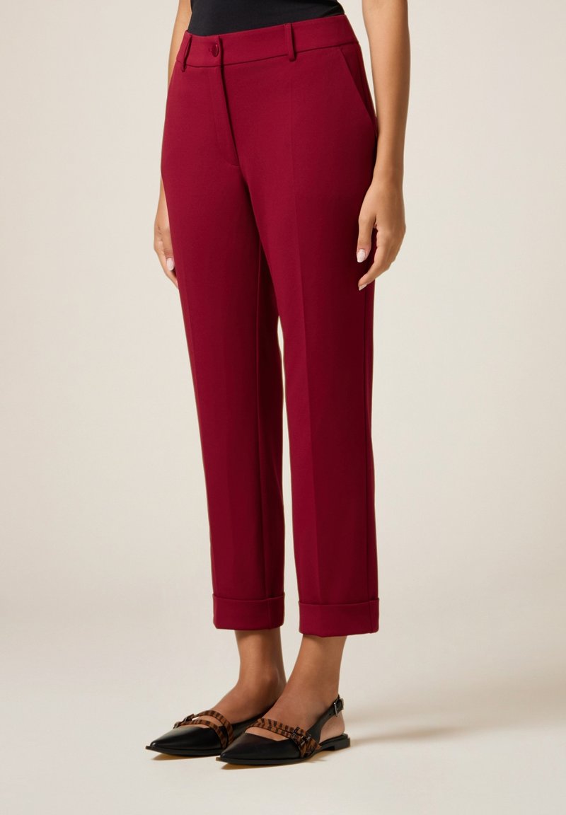 Kvinde iført bordeauxfarvede skræddersyede cropped bukser med ophøjede kanter og sorte spidse slingback flade sko med brune stribede stropper.