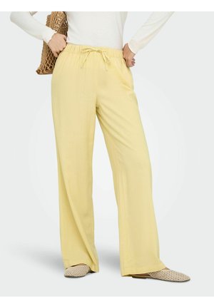 Žena má na sebe voľné svetložlté nohavice so šnúrkou, biely dlhý rukáv, tkanú tašku a béžové tkané slip-on topánky, stojí na sivom podlahe.