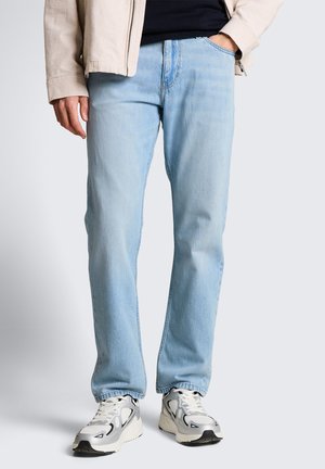 Mężczyzna w jasnoniebieskich dżinsach, białych i szarych trampkach, beżowej kurtce i czarnej koszulce, stojący na jednolitym szarym tle.