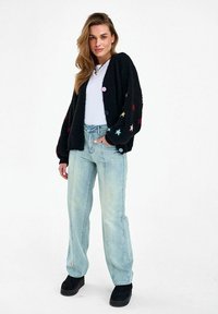 Žena v svetlomodrých rifle s vysokým pásom, bielej zatlačenej košeli, čiernom kardigáne s farebnými hviezdami a čiernych platformových topánkach.