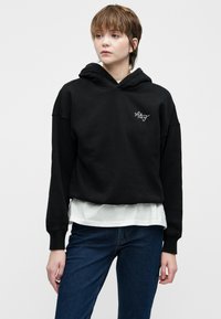 Noor inimene lühikese pruuni juustega, kandes musta lühendatud kapuutsiga jopperit, millel on "A&F" logo, valget särki alla, ja siniseid teksaseid, seismas valge tausta ees.