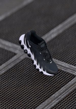 Mustavalkoinen Nike:n juoksukengät, joissa on erottuvat pyöristetyt pohjapolsterit, lepäävät tummalla metalliristikko pinnalla.