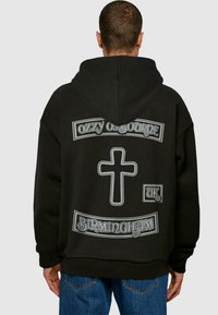 Sort hættetrøje med hvide grafikdetaljer: "OZZY OSBOURNE," et kors, "UK," og "BIRMINGHAM." Løs pasform og blød stofstruktur.