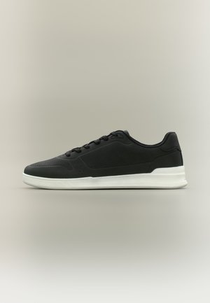 Baskets noires basses avec détails perforés, lacets noirs et semelle blanche épaisse sur un fond gris neutre.