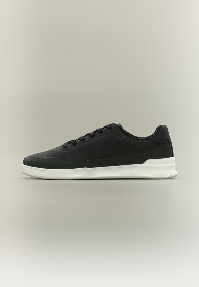 Baskets noires basses avec détails perforés, lacets noirs et semelle blanche épaisse sur un fond gris neutre.