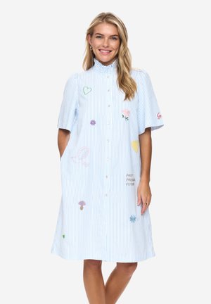 Smilende kvinde med langt blondt hår iført en lyseblå, knælængde kjole med striber og farverige broderede hjerter, blomster og en fugl.