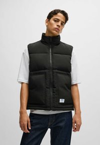 Black padded vest with high collar, front zipper, and two large pockets. Worn over a white t-shirt, featuring a branded label at the bottom.
