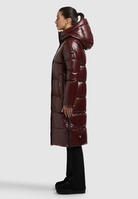 Bordó színű, fényes hatású, hosszú steppelt kabát kapucnival, oldalzsebekkel és cipzáras részletekkel. Fekete, bő szárú nadrággal és platform cipőkkel viselve.