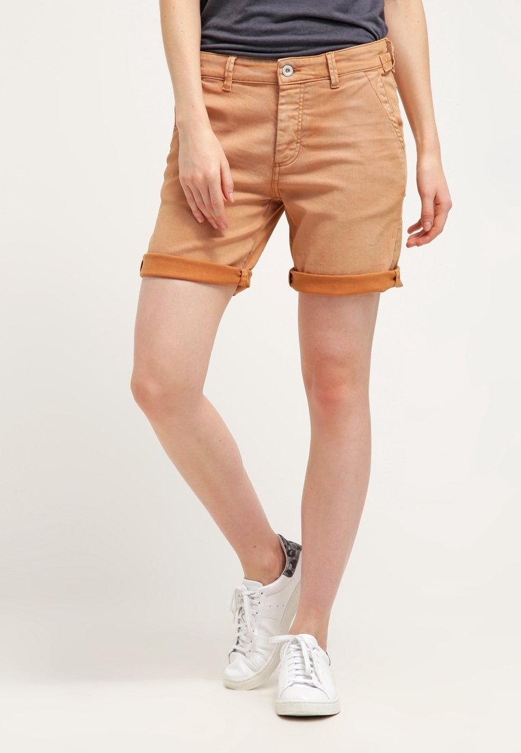 Shorts en denim beige à revers, design à cinq poches, fermeture à bouton sur le devant et texture légèrement décolorée. Portés avec des baskets blanches.