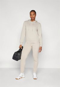 Beige sweatshirt med hætte og snore, parret med matchende tilspidsede bukser; holder en sort taske, iført hvide sko.