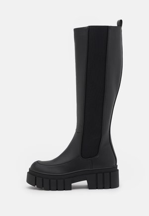 Botte haute en cuir noir avec une semelle épaisse et robuste et un panneau latéral élastique, conçue pour un enfilage facile et un style robuste.