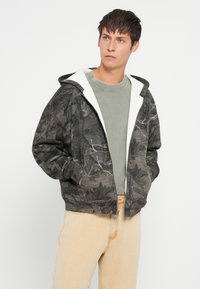 Padded mikina v tmavosivej farbe s vzorom listov, kapucňa s bielou fleecovou podšívkou, zips, bočné vrecká, nosená cez zelené tričko a béžové nohavice.