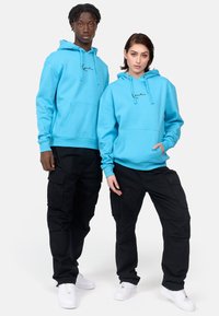 Two models wear matching hoodies in vibrant blue fleece, featuring a front pocket and drawstring hood, paired with black cargo pants and white sneakers.