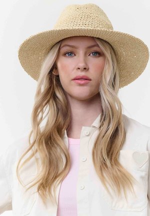 Blonde Frau mit langem, welligem Haar, die einen beigen, gewebten Hut mit breiter Krempe, ein helles, geknöpftes Hemd mit Herzdetail und ein pinkes Top vor weißem Hintergrund trägt.