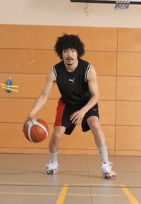 Καλαθοσφαιριστής με μαύρο αμάνικο μπλουζάκι και κόκκινες βερμούδες ντριμπλάρει μια μπάλα σε παρκέ γυμναστηρίου; φοράει γκρι και πολύχρωπα αθλητικά παπούτσια.