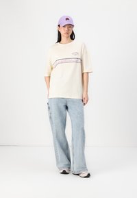 Beige ülepaisutatud T-särk graafilise bändiga, sinised laiad teksad ja helesinine kork brodeeritud kujundusega. Sneakerid mustade ja beežide aktsentidega.