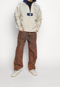 Ljus beige pulloverjacka med marinblå accenter och dragkedja, tillsammans med bruna texturerade oversized jeans och vita sneakers.