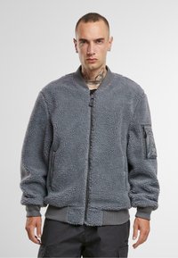 Giacca bomber in pile grigia con zip frontale, polsini a costine e tasca sul petto inclinata. Texture morbida e materiale peloso con vestibilità comoda.