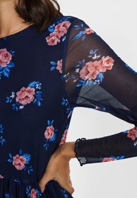 Vestido floral marino con rosas rosas y azules, mangas largas transparentes, tela texturizada, cintura fruncida y puños de mangas con volantes.
