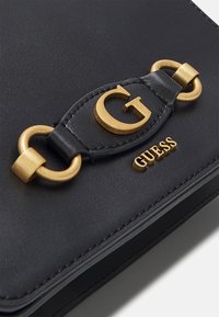 Černá kožená taška s kultovní zlatou kovovou „G“ logem a akcentem řetízku, s embosovaným nápisem „GUESS“ níže. Hladká textura.