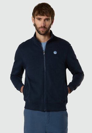 Man with beard wearing navy zip-up jacket and blue pants, standing with hands in pockets against plain gray background.