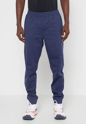 Granatowe dresy z elastyczną talią, zwężanymi nogawkami i teksturowanym wykończeniem materiału, połączone z białymi i pomarańczowymi butami sportowymi.