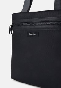 Schwarze Calvin Klein Tasche aus strukturiertem Material, mit einer vorderen Reißverschlusstasche und einem kleinen Logo-Tag. Die Träger sind aus Stoff mit matter Oberfläche.