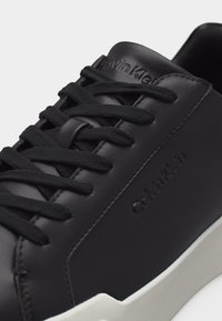 Μαύρο δερμάτινο αθλητικό παπούτσι Calvin Klein με μαύρα κορδόνια και λευκή σόλα, στενή λήψη που δείχνει το ανάγλυφο λογότυπο στο πλάι και στη γλώσσα του παπουτσιού.