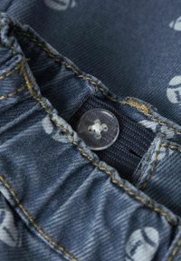 Zbliżenie na tkaninę dżinsową z marszczonym pasem, żółtym przeszyciem, przezroczystym guzikiem i nadrukowanymi elementami wzoru.