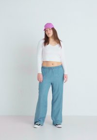 Biela dlhá rukávová crop top, svetlomodré nohavice na šnúrku, ružová čiapka, čierno-biele tenisky, stojí na neutrálnom pozadí.