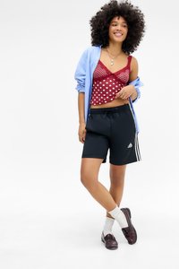 Young woman with curly hair wearing a light blue shirt over a red polka dot camisole, black Adidas shorts, white socks, and burgundy loafers.