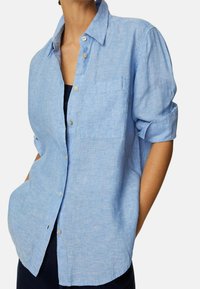 Camisa azul claro de botones hecha de lino. Presenta un bolsillo en el pecho, mangas arremangadas y un corte relajado con una superficie ligeramente texturizada.