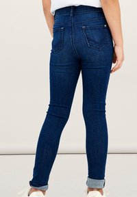 Jeans ajustados de mezclilla azul oscuro, con talle alto, bolsillos traseros y puños enrollados en los tobillos. Textura suave, diseño ajustado.