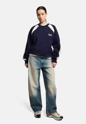 Młoda kobieta z ciemnymi włosami w niskim koku, nosząca duże kolczyki-koła, granatowo-białą bluzę, wyblakłe jeansy z szerokimi nogawkami i białe trampki.