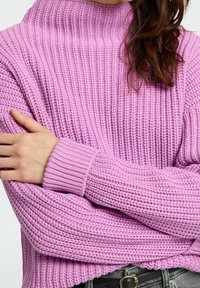 Pull-over en tricot rose pâle, avec un col montant, une texture côtelée et des manches larges. L'ourlet est légèrement arrondi, offrant une coupe décontractée.