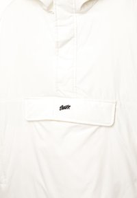 Weiße Jacke vorne mit einer mittigen Taschenklappe, die mit einem schwarzen, gestickten Markenlogo versehen ist, sowie einer vertikalen Naht über der Tasche.