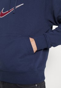 Osoba v tmavě modré mikině Nike s kapucí, s rukou v přední kapse, předvádějící vyšitý vínový a bílý logo Swoosh Nike.