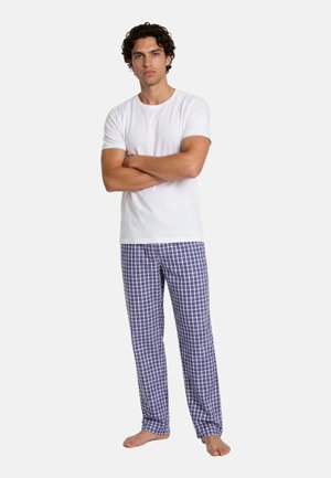 Man staat blootsvoets met gekruiste armen, draagt een effen wit T-shirt en blauw-wit geruite pyjamabroek.